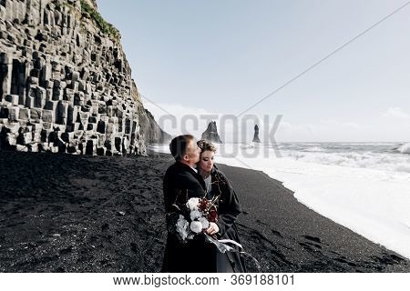 A Wedding Couple Walks Along The Sandy Black Beach Of Vik, Near The Basalt Rock, In The Form Of Pill