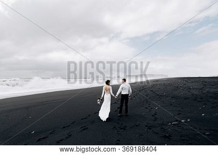 A Wedding Couple Is Walking Along The Black Beach Of Vic. Sandy Beach With Black Sand On The Shores 