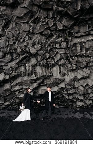 Destination Iceland Wedding. Wedding Couple Under A Rock Of Basalt Stones. Bride And Groom On The Bl