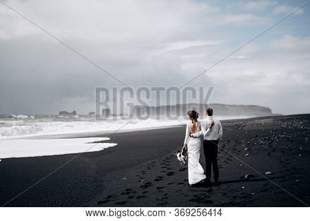 Destination Iceland Wedding. A Wedding Couple Is Walking Along The Black Beach Of Vic. Sandy Beach W
