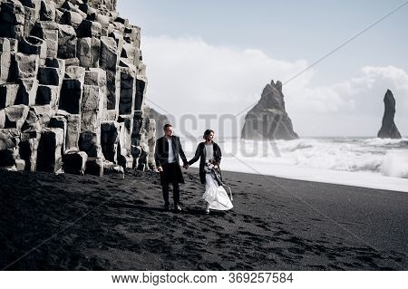 Destination Iceland Wedding. A Wedding Couple Walks Along The Sandy Black Beach Of Vik, Near The Bas
