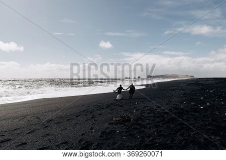 Destination Iceland Wedding. The Wedding Couple Runs Along The Sandy Black Beach Of Vik, Near The Ba