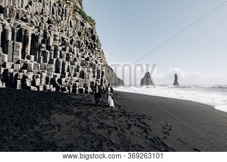 Destination Iceland Wedding. A Wedding Couple Walks Along The Sandy Black Beach Of Vik, Near The Bas