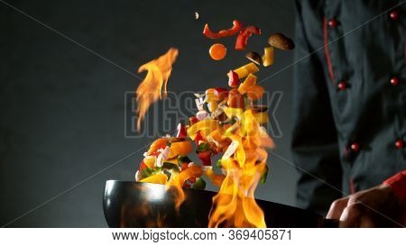 Closeup of chef throwing vegetable mix from wok pan in fire. Fresh asian food preparation on dark background.