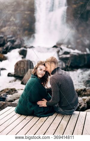 A Guy And A Girl Are Sitting Embracing On A Bridge Near Ehsaraurfoss Waterfall, Ehsarau River, Natio