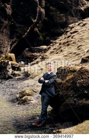 Portrait Of A Groom Leaning On A Stone In A Moss, Next To A Rocky Cleft With A Mountain River. Desti