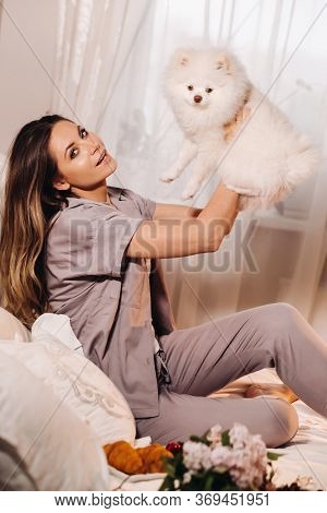 A Girl In Pajamas Sits In Bed At Night With Her White Dog Watching A Laptop And Eating Sweets.girl W