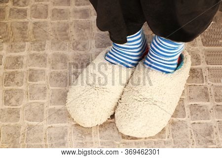 Feet In Fluffy Gray Wool Slippers On The Feet. Striped Fashionable Socks Are Worn On The Legs.
