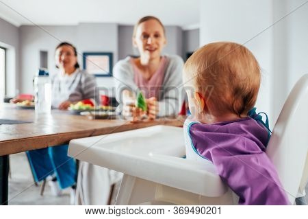 Young Mom Feeding Her Child With Solid Food In Dining Room. Cute Baby Girl Sitting On Highchair And 