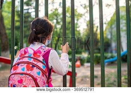 Little School Girl Sitting Next To School Fence Waiting For Going Back To Clases After Pandemic Outb