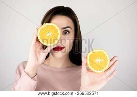 Young Brunette Woman With Red Lips Holding One Half Of Orange In One Hand And Another Half In Anothe