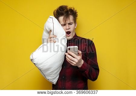 Sleepy Tired Guy Holds A Pillow And A Smartphone And Yawns In The Morning On A Yellow Isolated Backg