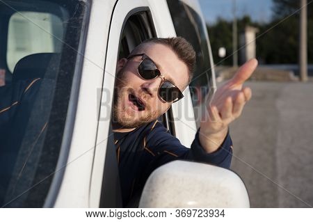 Young Man With Sunglasses Comes Out Of The Car Window With His Head With His Arm Raised. Face Expres