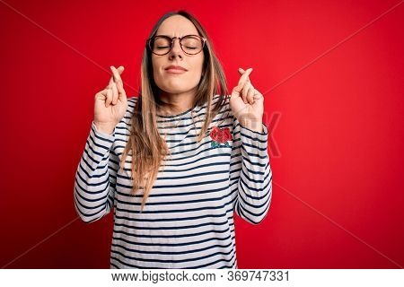 Young beautiful blonde woman with blue eyes wearing glasses standing over red background gesturing finger crossed smiling with hope and eyes closed. Luck and superstitious concept.