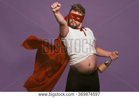 Drunk Super Antihero Holding Wine Bottle Showing His Tummy. Man In A Red Mask And A Superhero Cloak 