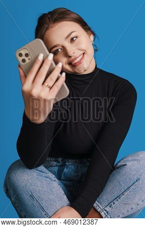 Excited Woman Using Mobile Phone Application Sitting On Chair Isoalted On Blue Studio Background. Te
