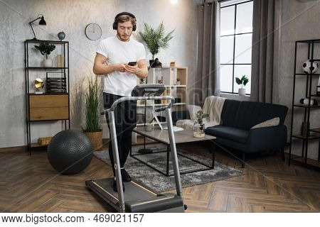 Pozitive Caucasian Man In Sports Clothes And Sneakers Listening Music In Headphones While Using Smar