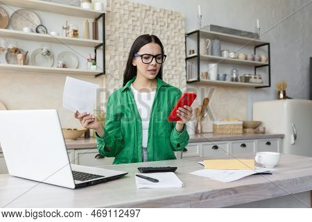 Angry Young Woman Sitting In The Kitchen At Home Using A Laptop. Holds Documents, A Letter In His Ha