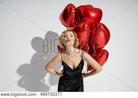Curly Blonde Woman In Black Slip Dress Pouting Lips Near Red Heart-shaped Balloons On Grey.