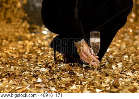 The Girl Picks Up A Glass Of Champagne From The Floor Amid A Pile Of Golden Confetti. Holiday Concep