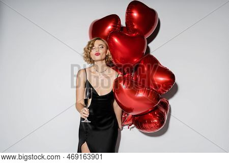 Curly Blonde Woman In Black Slip Dress Pouting Lips And Holding Glass Of Champagne Near Red Balloons