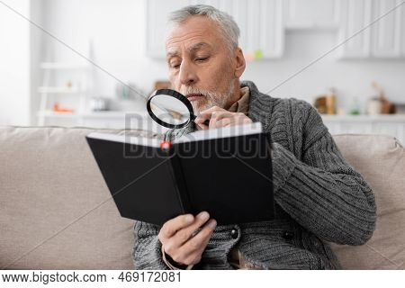 Senior Man With Azheimers Disease Looking In Notebook Through Magnifier While Sitting On Couch At Ho