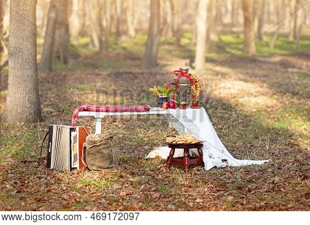 Samovar With Bagel, Candy On White Tablecloth. Sack, Accordion, Chair On Grass In Forest. Shrovetide