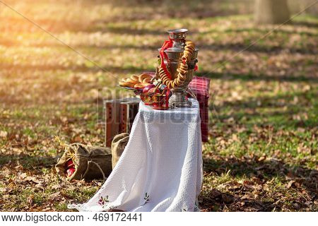 Samovar With Bagel On White Tablecloth. Side View. Sack On Grass In Forest. Shrovetide, Folk Festiva