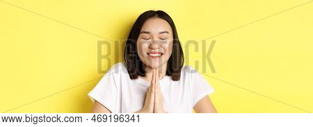 Close Up Of Hopeful Asian Girl In White T-shirt, Holding Hands In Pray, Namste Gesture And Smiling, 