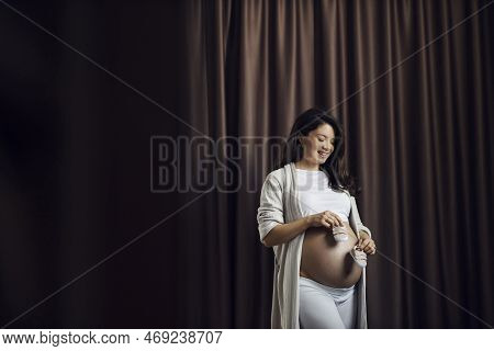 A Happy Pregnant Woman Is Standing With Her Naked Belly With Stretchmarks With Baby Shoes In Her Han