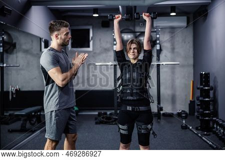 Woman Dressed In An Electric Muscular Suit Lifts The Dumbbells Above Her Head At The Training Center