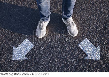 Choice Of Way. Man Standing In Front Of Drawn Marks On Road, Closeup. White Arrows Pointing In Diffe