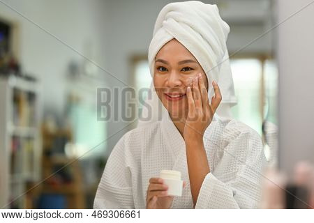 Smiling Beautiful Woman With Towel On Head Applying Nourishing Cream On Her Face And Enjoying Facial