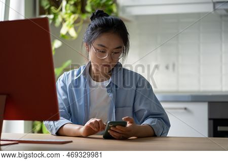 Young Asian Female Freelancer Scrolling Her Phone While Sitting At Desk In Front Of Computer. Remote