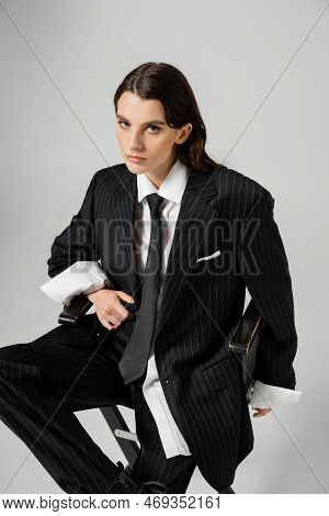 Brunette Woman Looking At Camera While Sitting In Black Stylish Pantsuit Isolated On Grey.
