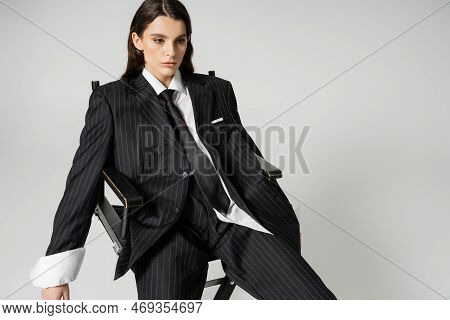 Brunette Woman In Black Striped Pantsuit And Tie Sitting On Chair Isolated On Grey.
