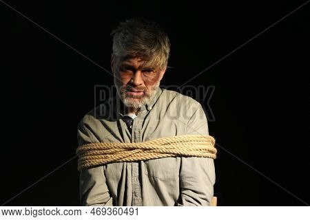 Beaten Man Tied With Rope On Black Background. Hostage Taking