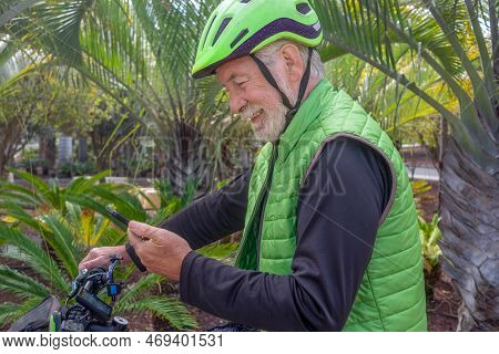 Smiling Senior Man Using Smart Phone While Bike Ride In Outdoor Tropical Place. White-haired Retiree