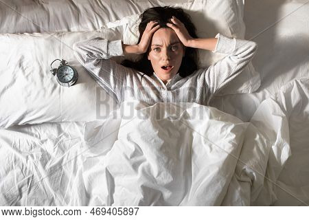 Shocked Young European Woman In Pajamas Woke Up, Holding Her Head, Lies On White Bed With Alarm Cloc