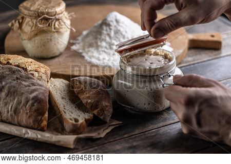 Male Hands Open A Jar With Active Yeast, Fresh Bread And Pastries In The Background.