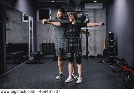 Young Woman Workout In Electric Muscular Suit, Stimulate Exercise At Training Center. A Male Trainer