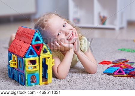Cute Curly Girl Next To A House Made Of A Magnetic Constructor At Home On The Carpet In The Children