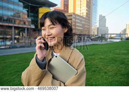 Stylish Smiling Girl, University Student In Trench Coat, Holds Tablet, Talks On Mobile Phone, Has Co