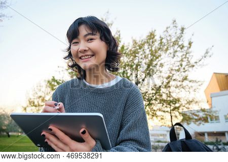 Close Up Portrait Of Young Korean Girl Sits Outdoors In Park, Holds Her Digital Tablet And Graphic P