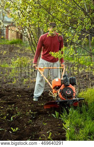 A Farmer Man Works In The Field, Plowing The Land With A Plow On The Farm. A Plowman On A Walk Behin