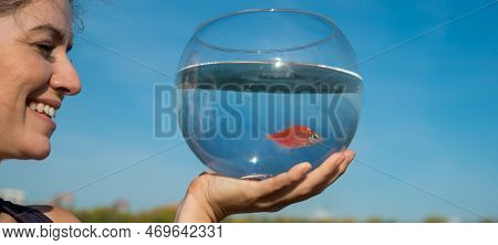 Woman Holding Round Aquarium With Goldfish On Blue Sky Background.