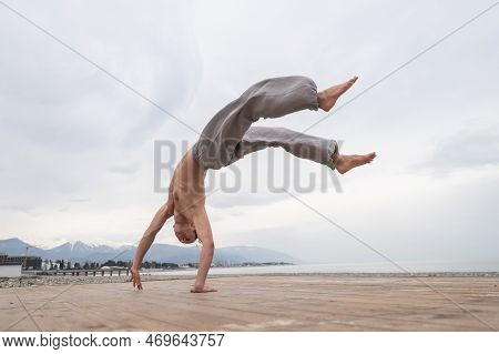 Shirtless Caucasian Man Doing Backflip On Pebble Beach.