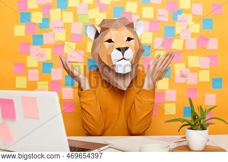 Indoor Shot Of Helpless Confused Man In Lion Mask Wearing Orange Sweater Sitting At Workplace With L