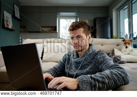 Man Using Laptop In Living Room, Dog Lying On Sofa At Background. Freelancer Working At Home Workpla