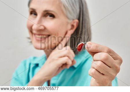 Senior Woman Holding Hearing Aid In Hand On Foreground And Pointing Finger At Her Ear, Soft Focus. T